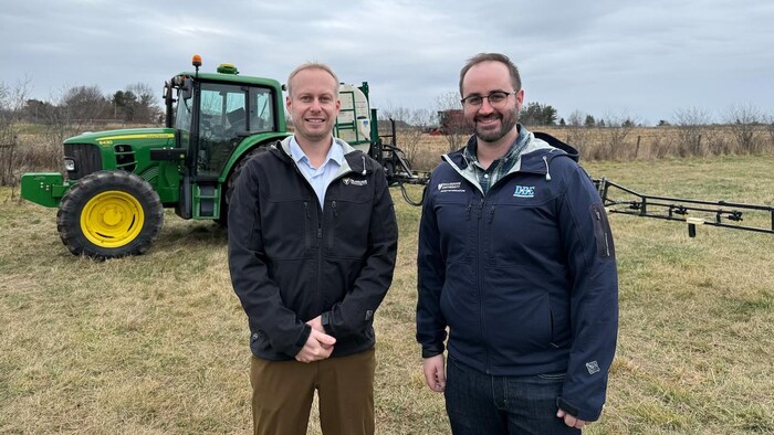 Deux hommes posent pour une photo dans un champ devant un tracteur vert et jaune, sous un ciel voilé.