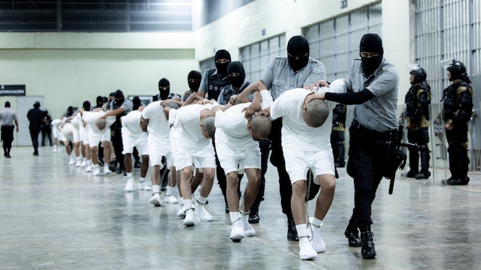 Dans une salle, des policiers tiennent des prisonniers habillés en blanc.