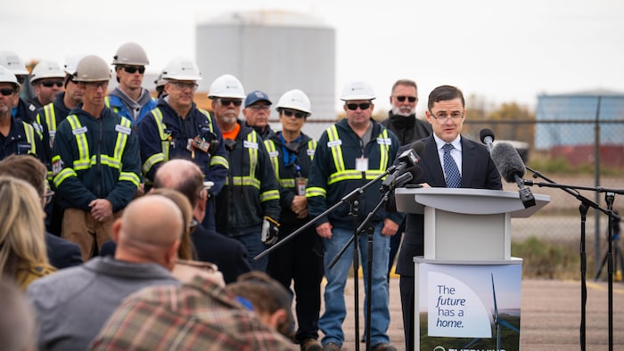 Un homme en complet aux cheveux gominés peignés vers la gauche, portant ses lunettes sur le bout de son nez, est debout devant des micros. Derrière lui, des travailleurs de la construction en uniforme, casqués.