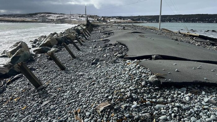 De gros morceaux d'asphalte arrachés et des roches sur une route détruite au-dessus d'un cours d'eau, en hiver.