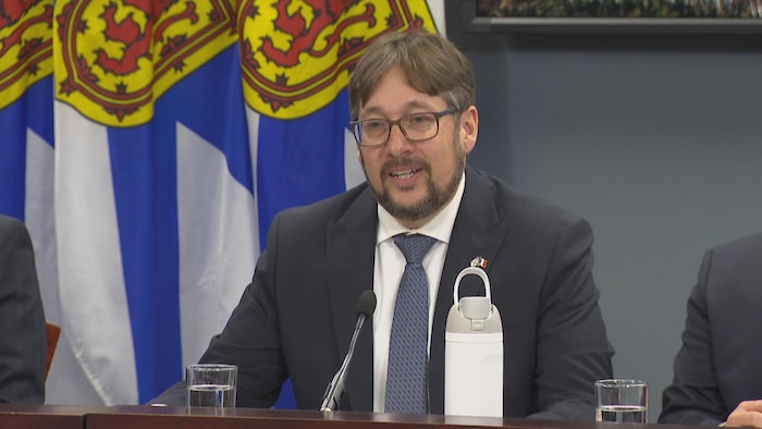Un homme est assis à une table devant des drapeaux de la Nouvelle-Écosse.