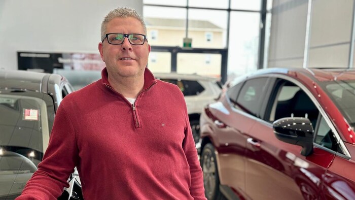 Un homme accoudé sur le capot d'une voiture noire reluisante dans la salle de montre d'un concessionnaire automobile bien éclairé.