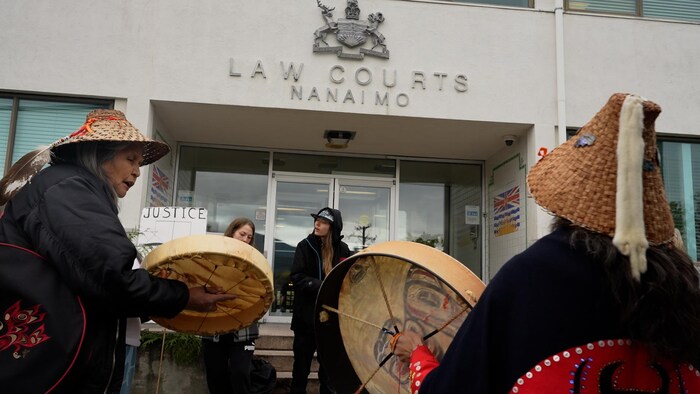Des personnes devant le tribunal de Nanaimo jouent du tambour, certaines vêtues de chapeaux de cèdre.