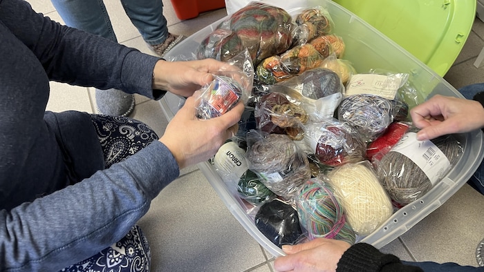 Deux personnes manipulent des pelotes de laine multicolores rangées dans un grand bac en plastique transparent. La plupart des écheveaux sont emballés individuellement dans des sachets protecteurs. On aperçoit des marques comme Caron et Jubilée parmi le stock.
