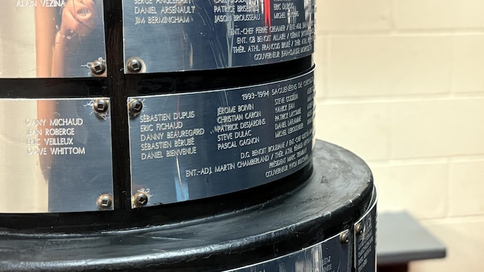 Un plan rapproché montre les plaques d'argent vissées sur la base d'un trophée de hockey, gravées de noms de joueurs et d'équipes. À droite, un arrière-plan flou révèle une bannière noire ornée du logo Proxi.