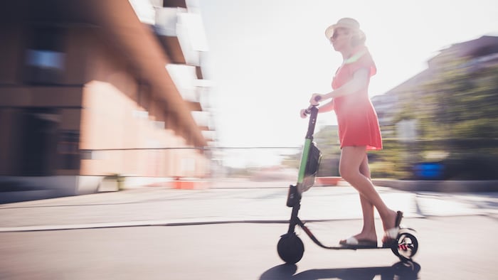 Une femme en robe estivale rouge, coiffée d'un chapeau de paille et portant des lunettes de soleil, roule à trottinette dans un paysage urbain, devant un bloc appartement.