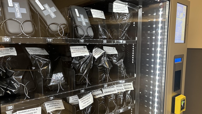 Un distributeur automatique en verre est rempli de produits de réduction des méfaits. Les articles sont emballés individuellement dans des sacs noirs ou des boîtes, étiquetés "NALOXONE" ou "TROUSSE NALOXONE & BANDELETTES À DÉTECTION DU FENTANYL". Les étagères sont éclairées, et un écran tactile ainsi qu'un bouton jaune sont visibles sur le côté droit de la machine.