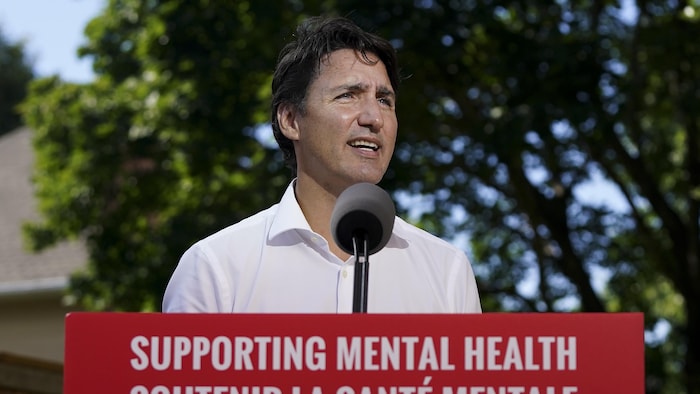 M. Trudeau parle aux médias, de face, à l'extérieur. Devant lui, on lit « Soutenir la santé mentale ». En arrière-plan, des arbres, une clôture et une maison de banlieue.