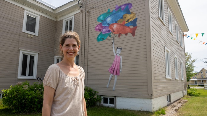 Catherine Rondeau sourit à la caméra devant une de ses oeuvres représantant une petite fille tenant un bouquet de ballons en forme de truites multicolores.