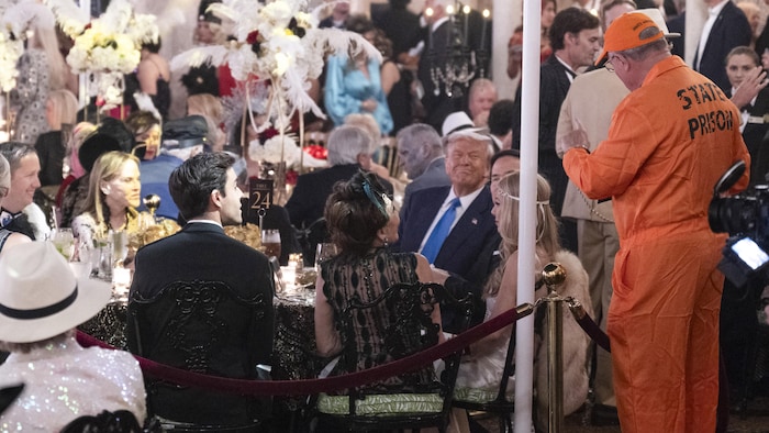 Donald Trump, assis à array avec des invités, souriant à un homme debout déguisé en prisonnier.