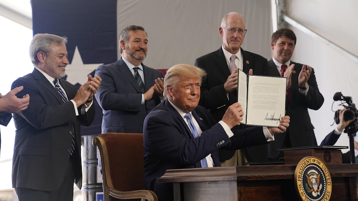 Donald Trump sourit à la caméra en tenant dans ses mains le permis qu'il vient de signer. Derrière lui, cinq hommes l'applaudissent.