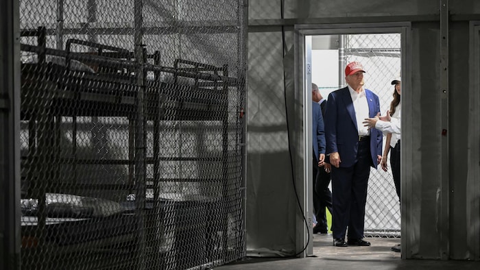 Donald Trump, portant sa classique casquette Make America Great Again rouge, se tient dans l'embrasure de la porte d'une cellule dans un centre de détention, où plusieurs lits sont superposés derrière une clôture en grillage.