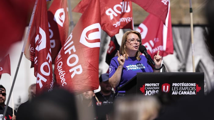 Une femme parle derrière un podium, devant de multiples drapeaux d'Unifor.