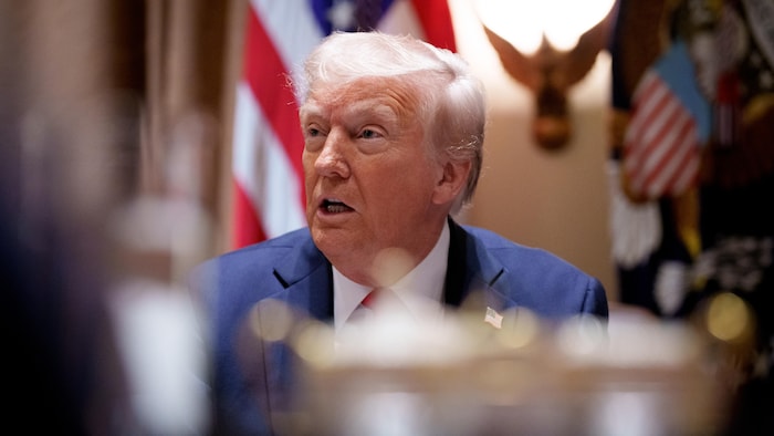 Donald Trump parle, assis devant un drapeau des États-Unis.