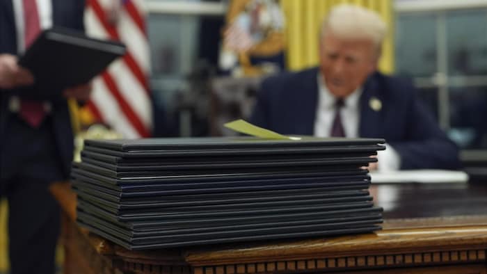 Une série de documents est sur le bureau ovale, avec Donald Trump en arrière-plan.