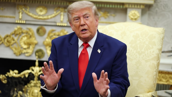 Donald Trump, vêtu d'un costume bleu et d'une cravate rouge, gesticule des mains en parlant, assis dans un fauteuil.
