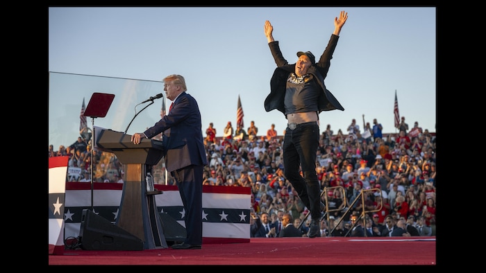 Donald Trump, les yeux fermés, parle au micro tandis qu'Elon Musk, les bras dans les airs, saute à ses côtés sur la scène d'un rassemblement extérieur.