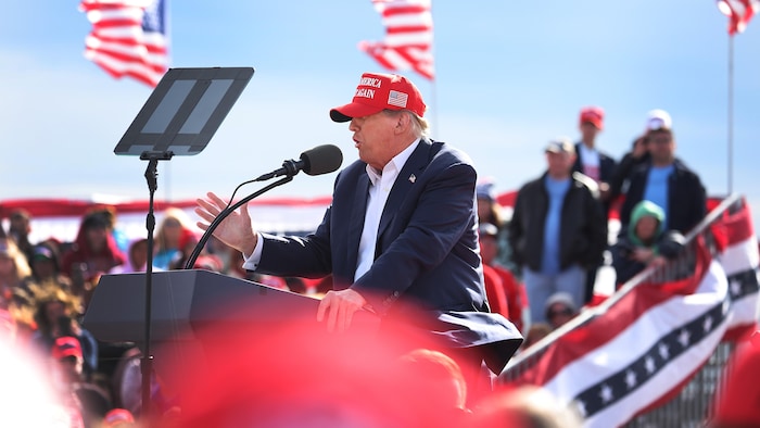 Donald Trump, à un lutrin, parle dans un micro pour s'adresser à ses partisans, avec en toile de fond des drapeaux américains.