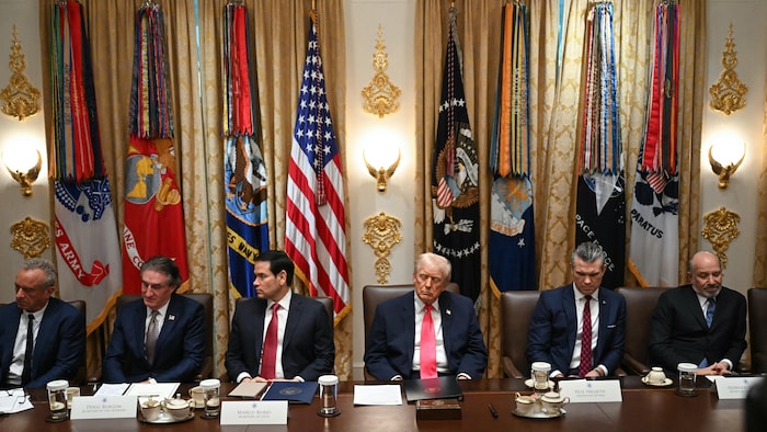Donald Trump est entouré de plusieurs de ses secrétaires dans la salle du Cabinet, où sont exhibés plusieurs drapeaux.
