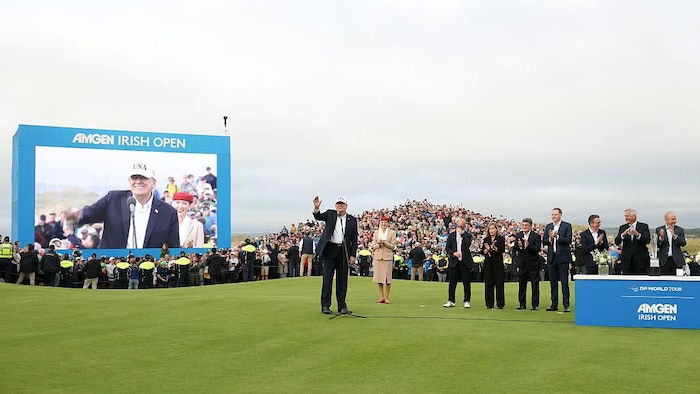 Donald Trump sur une pelouse de golf, en train de parler dans un micro, avec son image diffusée sur un écran, tandis que des gens l'applaudissent.