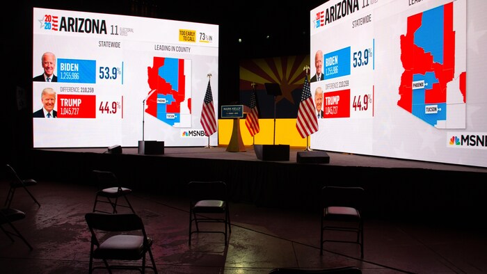 Deux écrans sur lesquels sont projetés les résultats de la soirée électorale, devant lesquels se trouvent plusieurs chaises vides.