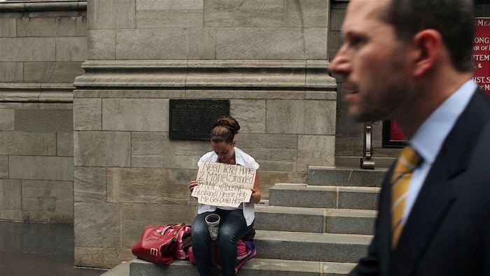 Un homme en complet passe devant une femme tenant un panneau pour demander de l'argent dans la rue
