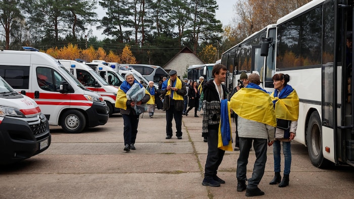Des personnes enveloppées dans des drapeaux ukrainiens se rassemblent près d'autobus, alors que des habitants évacués dans le cadre d'une mission humanitaire depuis la ville d'Oleshky, occupée par la Russie, regagnent le territoire contrôlé par l'Ukraine le 31 octobre 2026.