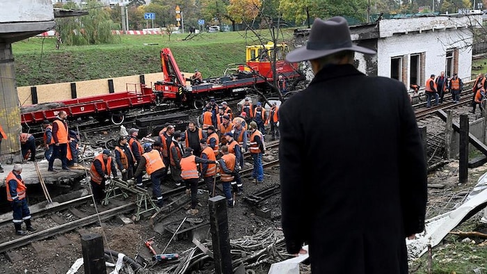 Un homme regarde des travailleurs vêtus d'un gilet orange s'affairer sur des rails où des traces de destruction sont visibles.