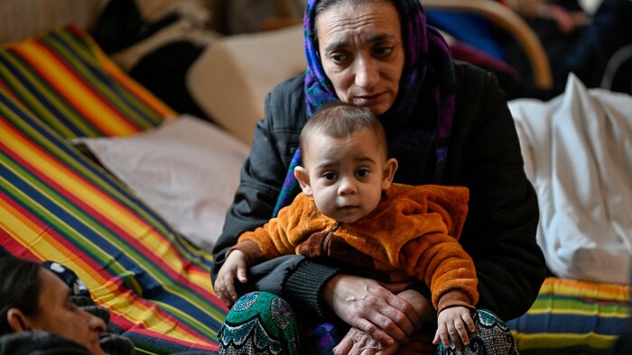 Une réfugiée, assise sur un lit de fortune, tient un enfant sur ses genoux.