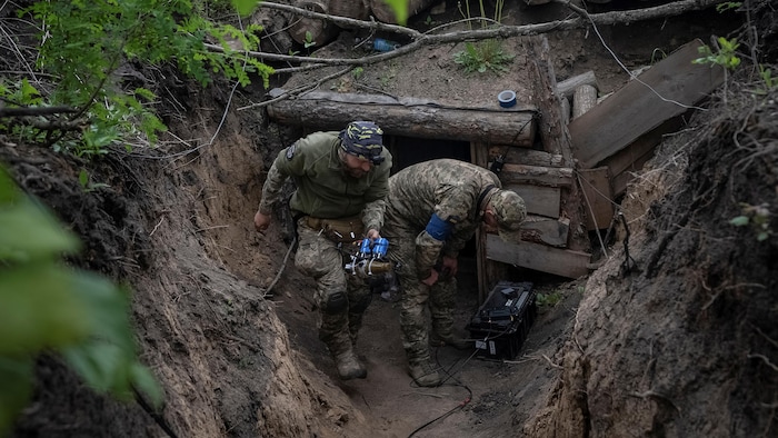 Two Ukrainian soldiers lie in a ditch, one carrying a drone.