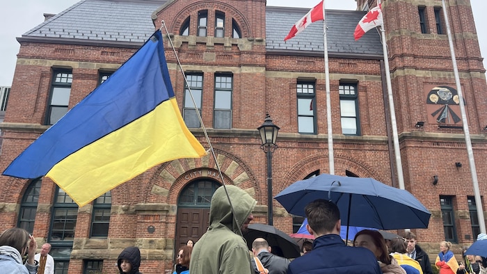 Un groupe de personnes devant l'hôtel de ville. Un homme tient un grand drapeau de l'Ukraine. Un autre a un parapluie bleu.
