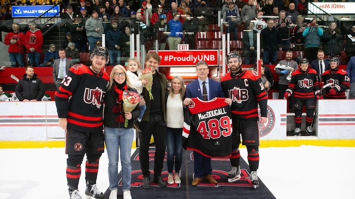 L'entraîneur pose avec sa famille et deux joueurs, debout sur un tapis déroulé sur la patinoire. Il exhibe un chandail de hockey avec son nom et le numéro 489.