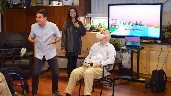 Des gens devant un écran avec un joueur, plus âgé, muni d'un casque de réalité virtuelle.