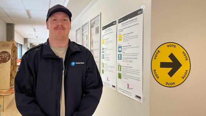Simon Plourde se tient debout devant une affiche qui indique la salle où voter. 