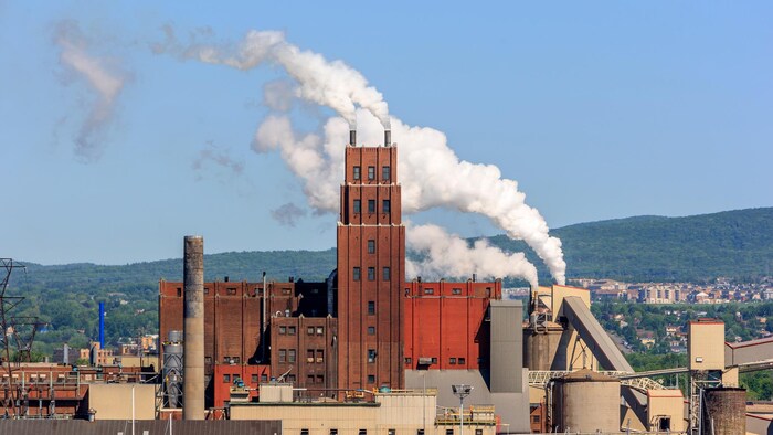Une usine dont les cheminées dégagent de la fumée. Au loin, on aperçoit la ville.