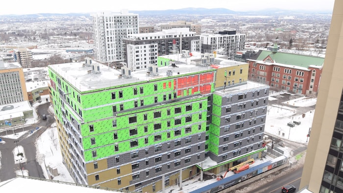 Un immeuble en construction vu en plongée, d'un autre immeuble. À l'arrière-plan, la Basse-Ville de Québec et des montagnes de la chaîne des Laurentides. 