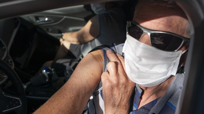 Un homme dans sa voiture et portant un masque montre l'endroit sur son épaule où il s'est fait vacciner.