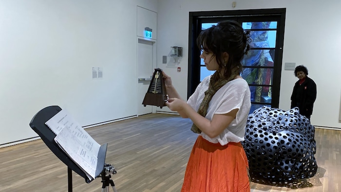 Una mujer pone a funcionar un metrónomo en una sala de museo.  · Une femme fait marcher un métronome dans un salle de musée. 