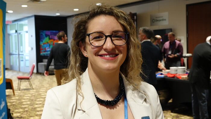 Une jeune femme avec des lunettes sourit à l'objectif, elle est proche de plusieurs kiosques.