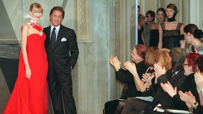 Devant un public qui applaudit, Valentino pose avec Claudia Schiffer, qui porte une longue robe rouge, lors d'un défilé. 