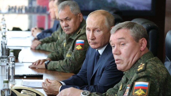 Vladimir Poutine au centre, en costume et cravate, assis à une table lors d'une réunion; Sergueï Choïgou et Valéri Guerassimov en uniforme militaire. 