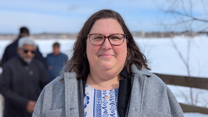 Une femme souriante dehors en hiver devant le fleuve. On distingue quelques personnes floues à l'arrière.