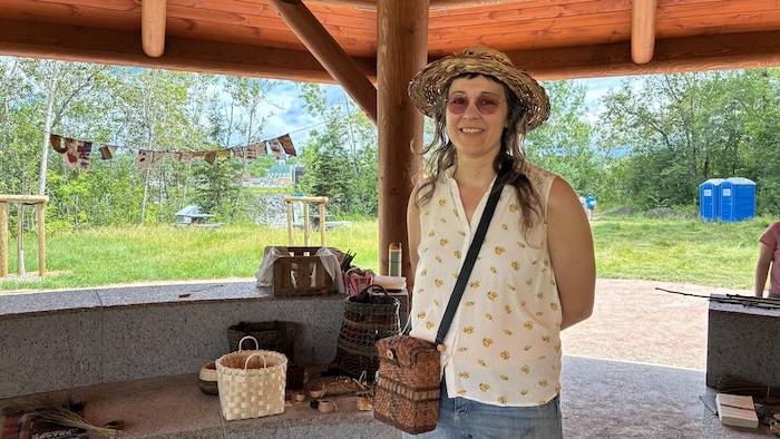 Une femme souriante portant un chapeau de paille, des lunettes teintées et une camisole à motifs pose sous une structure en bois. Près d'elle, divers paniers artisanaux sont exposés sur un muret en pierre, avec la nature en arrière-plan.