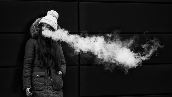 Une jeune femme adossée à un mur vapote. Elle porte une tuque et un manteau d'hiver.