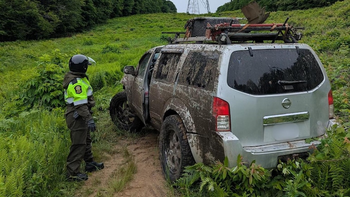 Un policier s'apprête à parler au conducteur qui entrouvre la portière d'une camionnette salie de boue sur tout un côté, sur un terrain boueux sous des lignes à haute tension.
