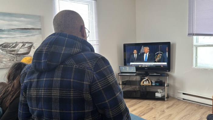 Vu de dos, un homme vêtu d'un manteau à carreaux bleus regarde une diffusion télévisée montrant Donald Trump. La pièce lumineuse comprend une peinture de paysage marin au mur et un ordinateur portable posé sous l'écran de télévision.