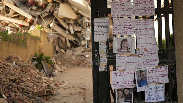 Des avis de recherche collés sur un mur avec des décombres du séisme en arrière plan.