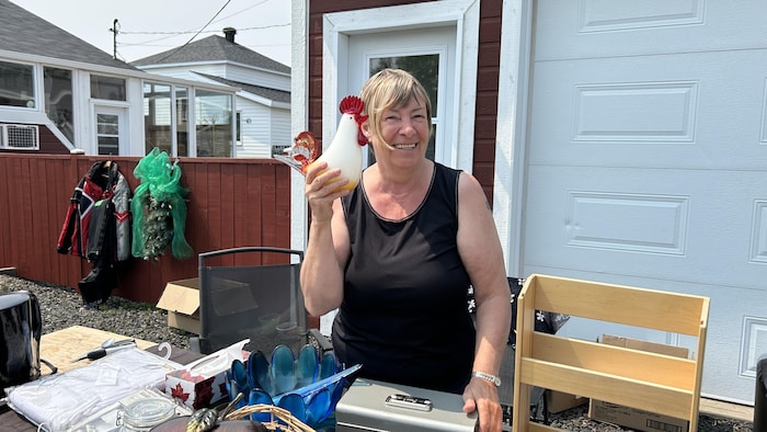 Louise Raymond sourit devant une table d'objets à vendre. Elle tient dans sa main près de son visage, une sculpture en verre représentant un coq.