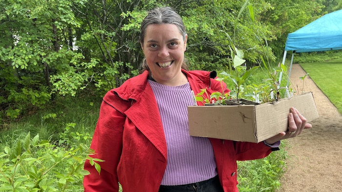 Une femme dehors devant un arbre, tenant une boîte remplie de plantes naissantes en pot. Elle regarde la caméra en faisant un grand sourire.