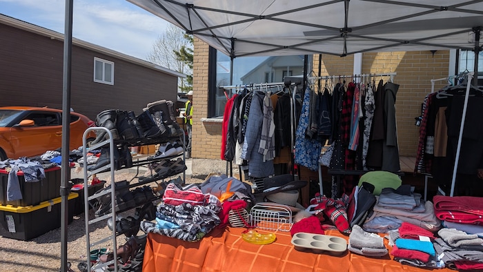 Une table, des portants de vêtements et des étagères de chaussures sont installés sous un auvent de vente de garage.